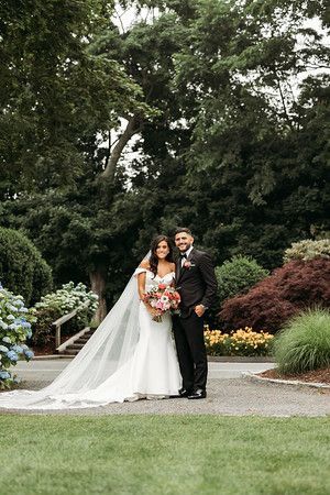 Bride and groom posing outdoors, smiling. Bride in white dress, veil, holding bouquet; groom in black suit.