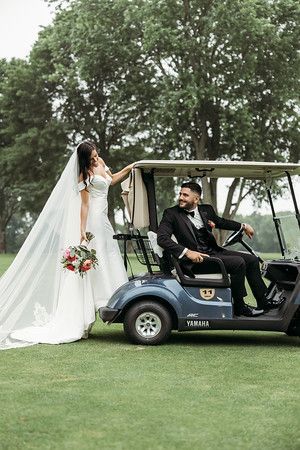 Bride and groom posing by a blue golf cart on a green golf course. The bride wears a white gown and veil, holding flowers. The groom is in a suit.