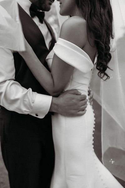 Couple embracing, groom in tuxedo, bride in white off-the-shoulder dress. Outdoor setting, black and white photo.