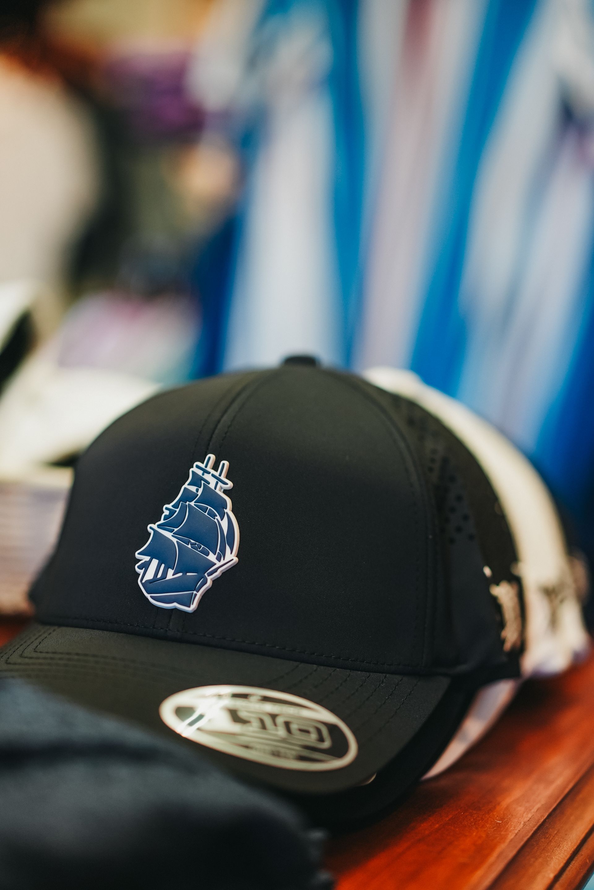 Black baseball cap with blue ship logo, stacked with white and black hats.