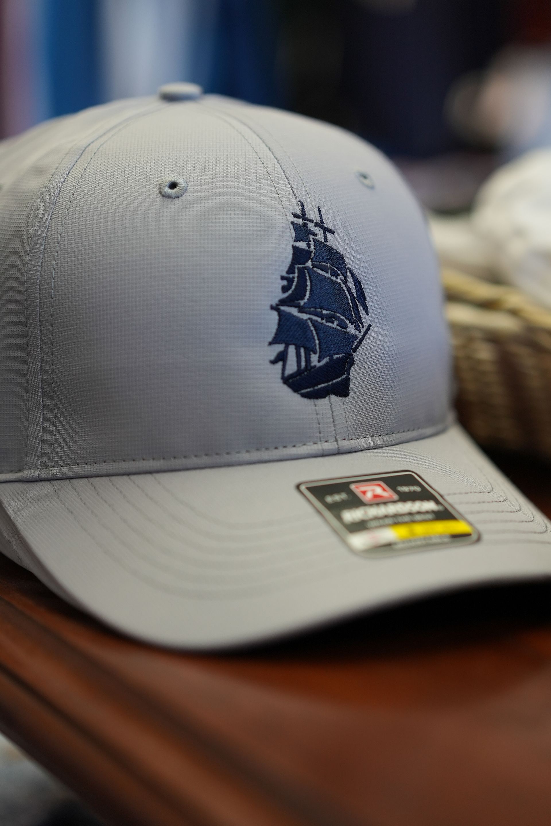 Gray baseball cap with dark blue embroidered sailboat; sits on a brown surface.