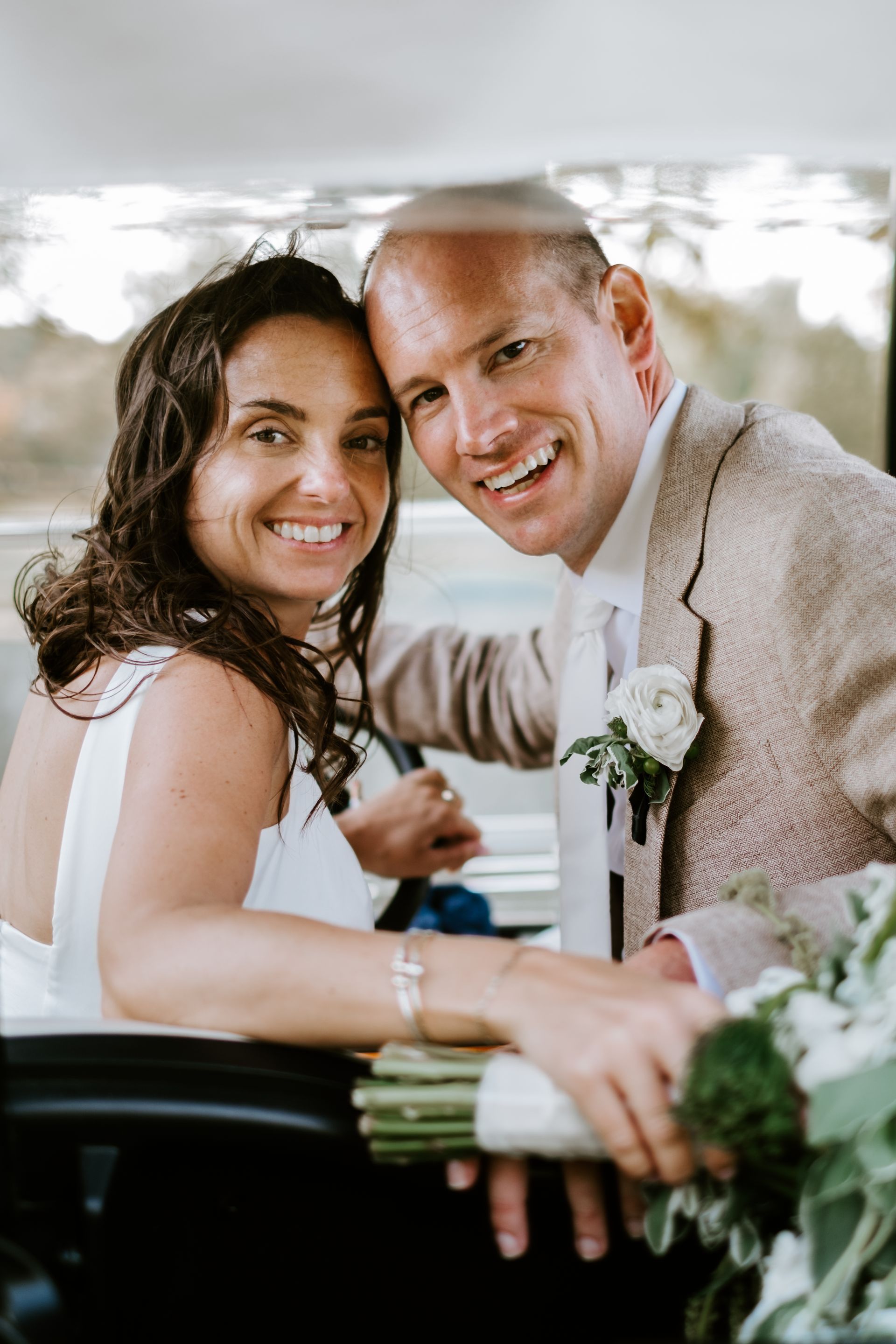 Newlyweds smiling in a golf cart. Bride in white dress, groom in tan jacket. Green bouquet.