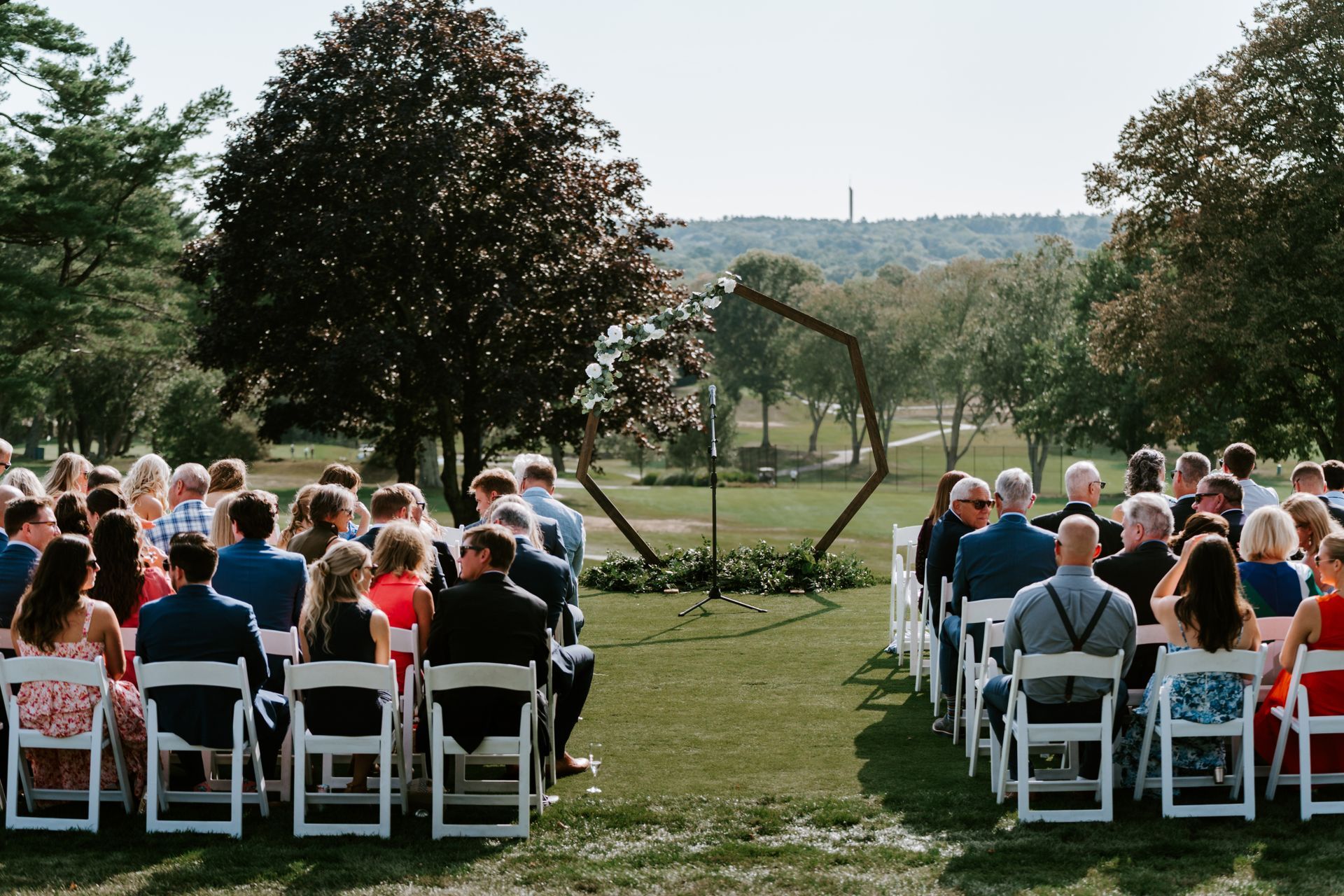 Wedding ceremony: Guests seated, facing a geometric arch, outdoors on a sunny day.