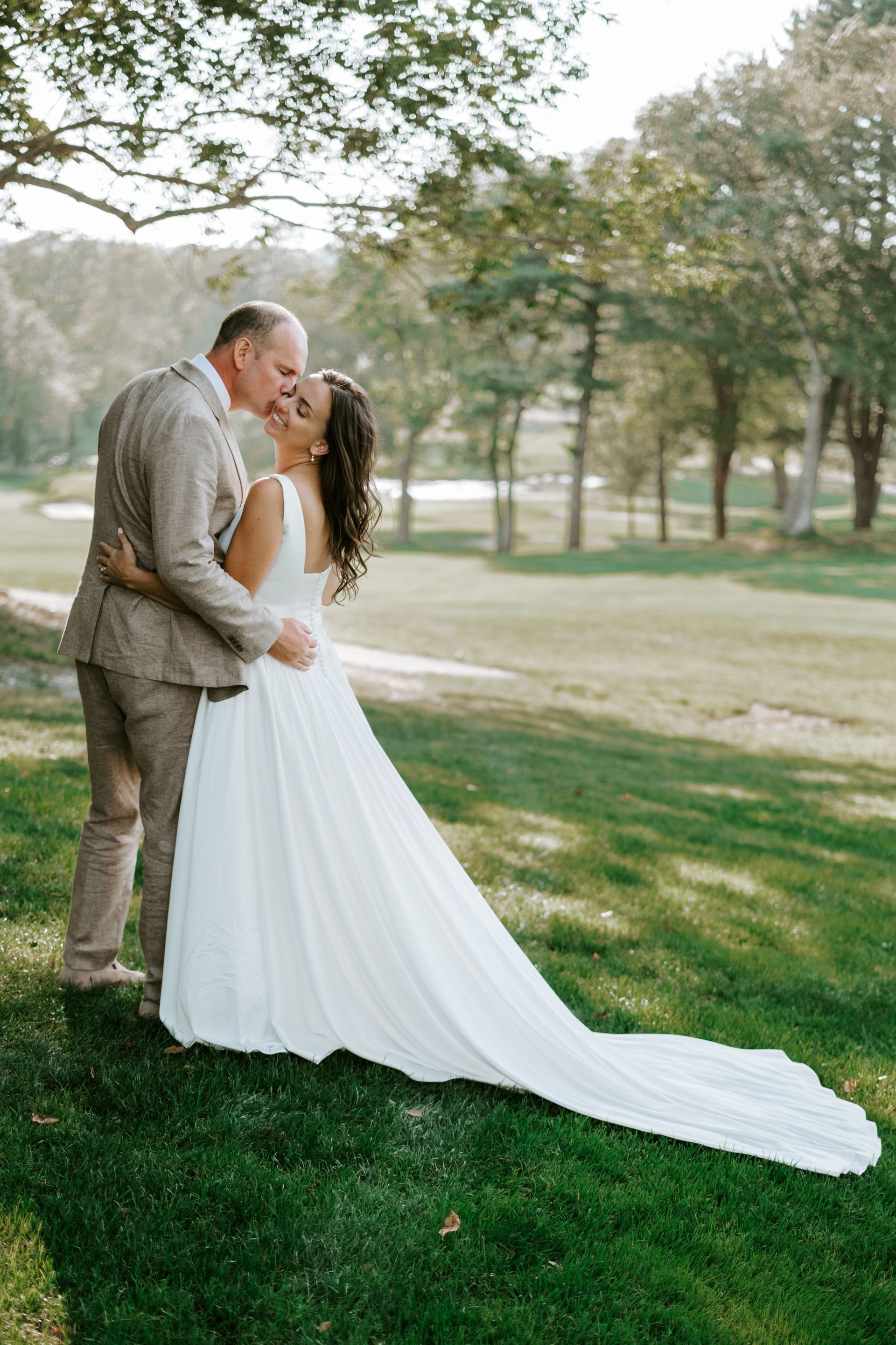 Bride and groom kissing on a golf course. She wears a white dress with a long train. He wears a tan suit.