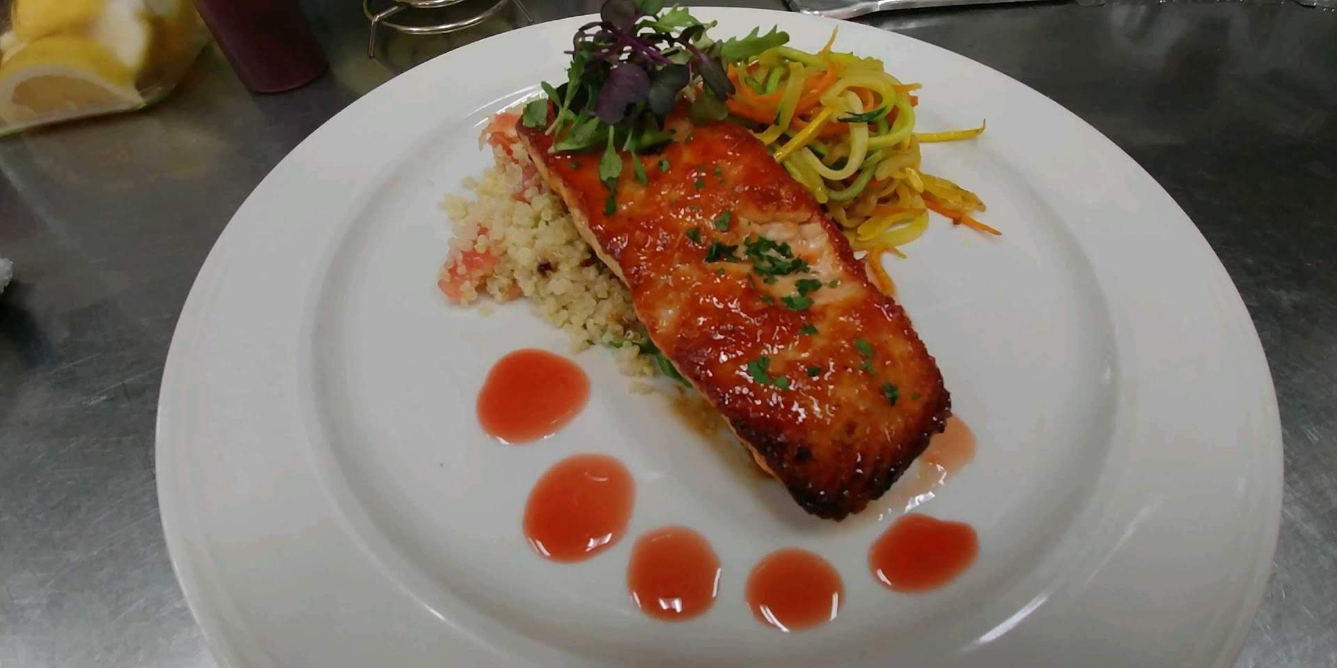 Salmon fillet with sauce and greens on a white plate, garnished with colorful vegetable noodles and sauce droplets.