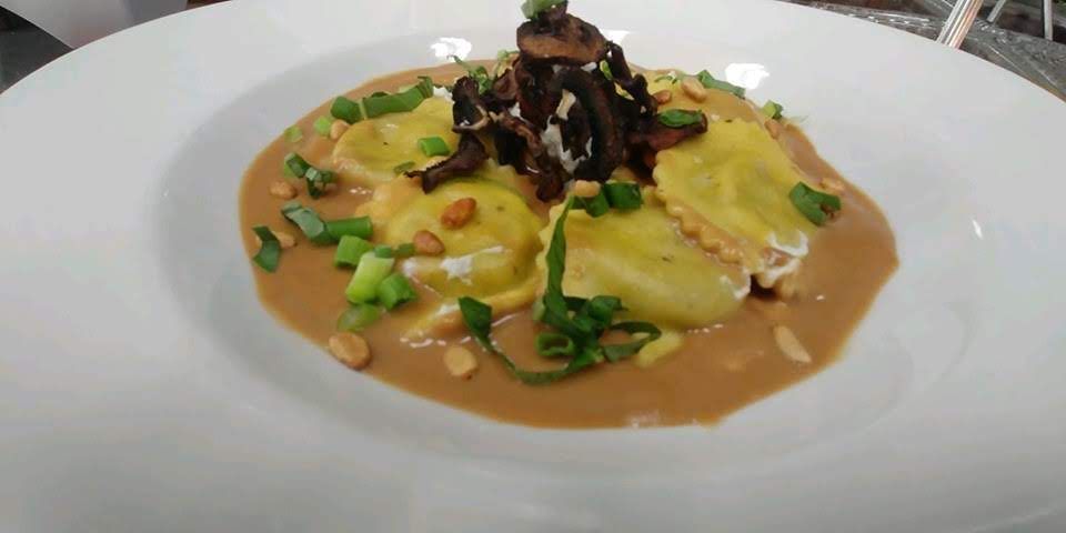 Ravioli dish with brown sauce, mushrooms, green onions, and pine nuts on a white plate.