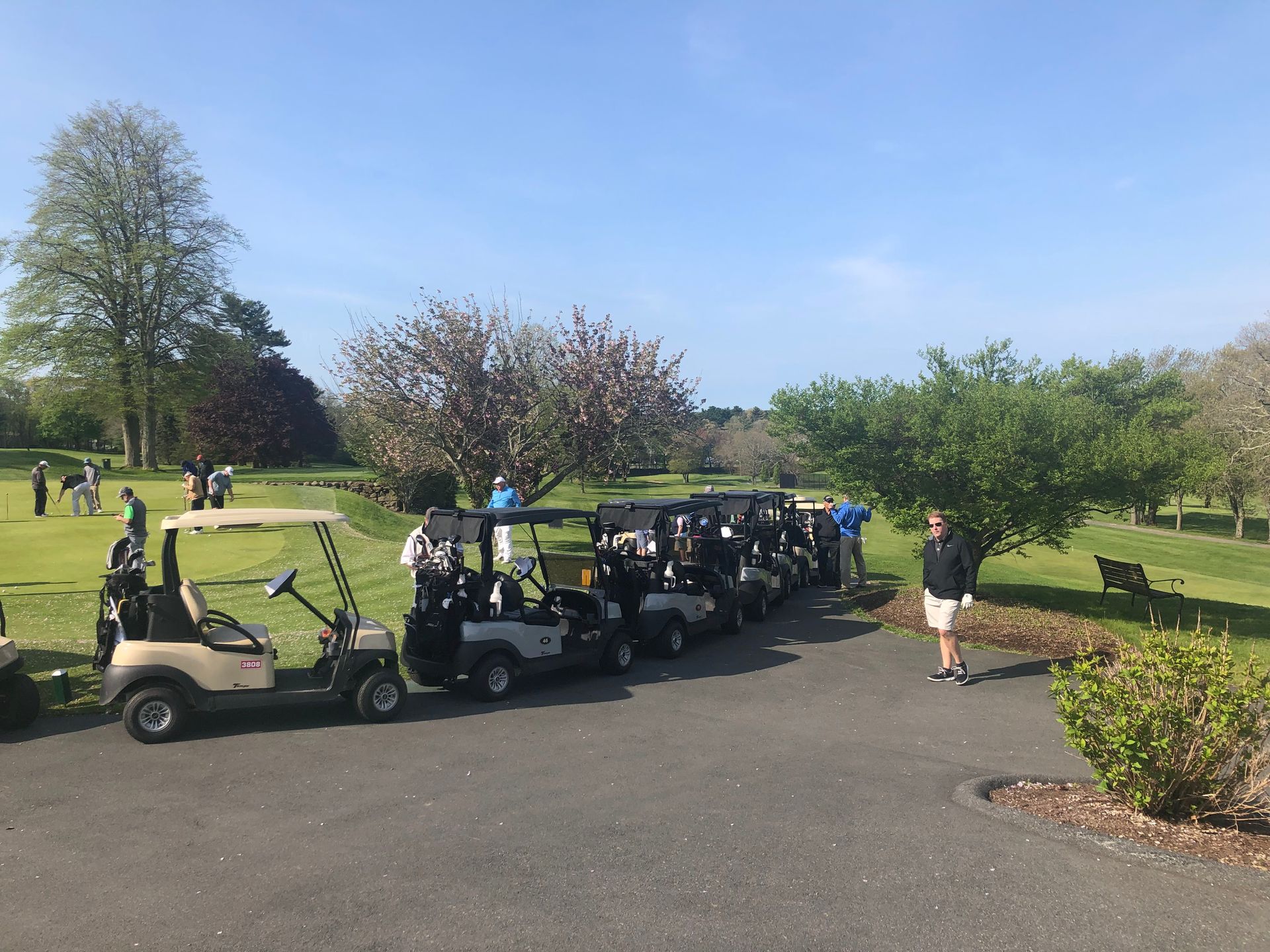 Golf carts lined up on a paved path next to a green golf course. People are visible nearby.