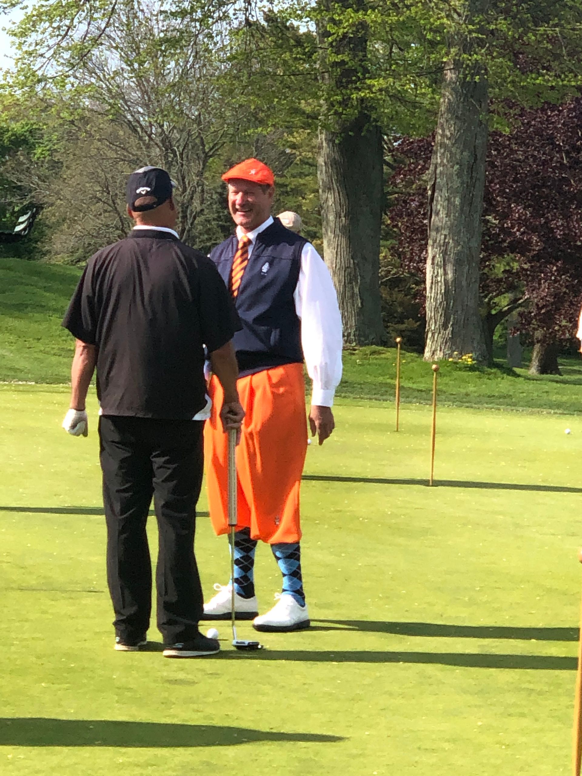 Two men on a golf course. One wears orange pants, hat, and patterned socks; the other, black.