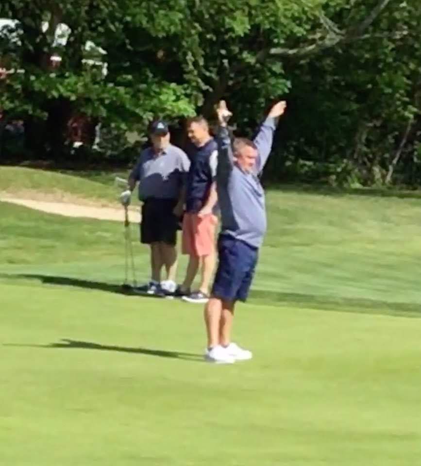 Man celebrates on a golf course, arms raised. Two men watch on a green lawn with trees in the background.