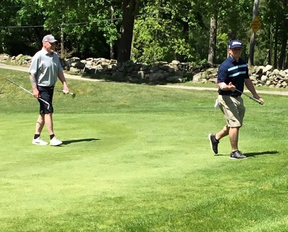 Two golfers on a green field, holding clubs. Sunny day, with trees and a stone wall in the background.
