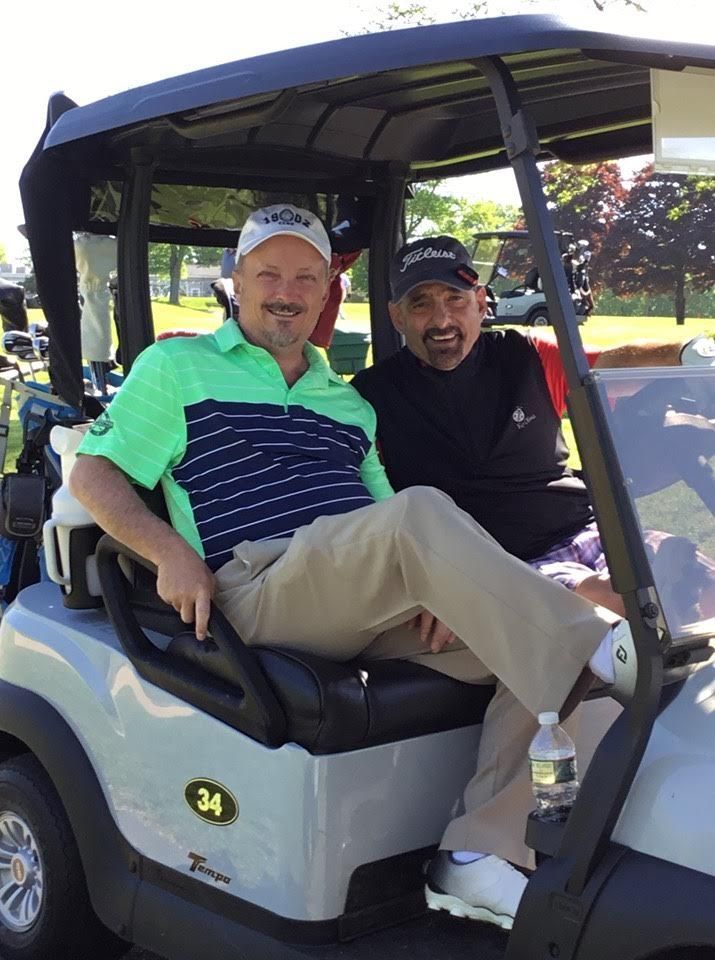 Two men smiling, sitting in a golf cart on a sunny day. One wears a golf shirt, the other a black shirt.