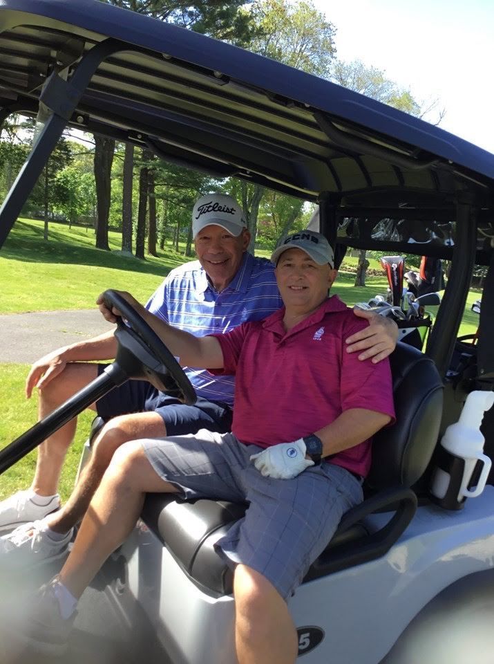 Two men in golf cart, smiling. One driving, the other has arm around him, sunny outdoors.