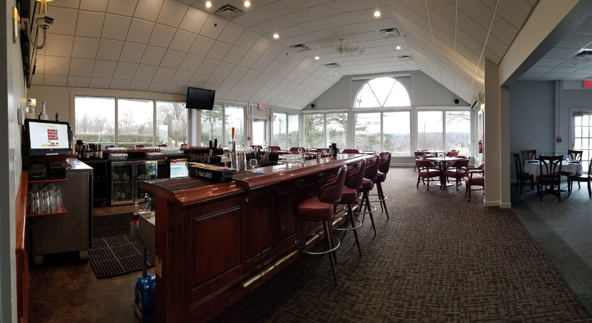 Interior view of a bar with a curved wooden counter, burgundy stools, and tables. Large windows provide exterior views.