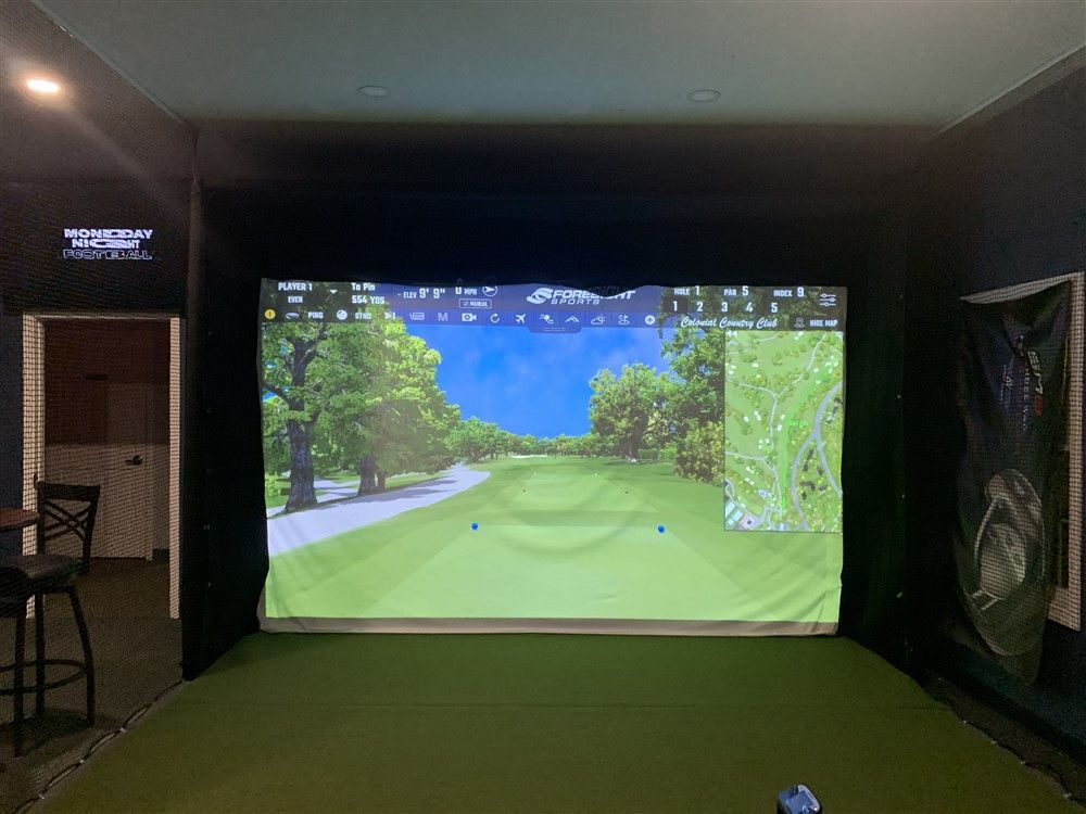 Golf simulator setup with screen displaying a golf course view. Green turf floor and black walls.
