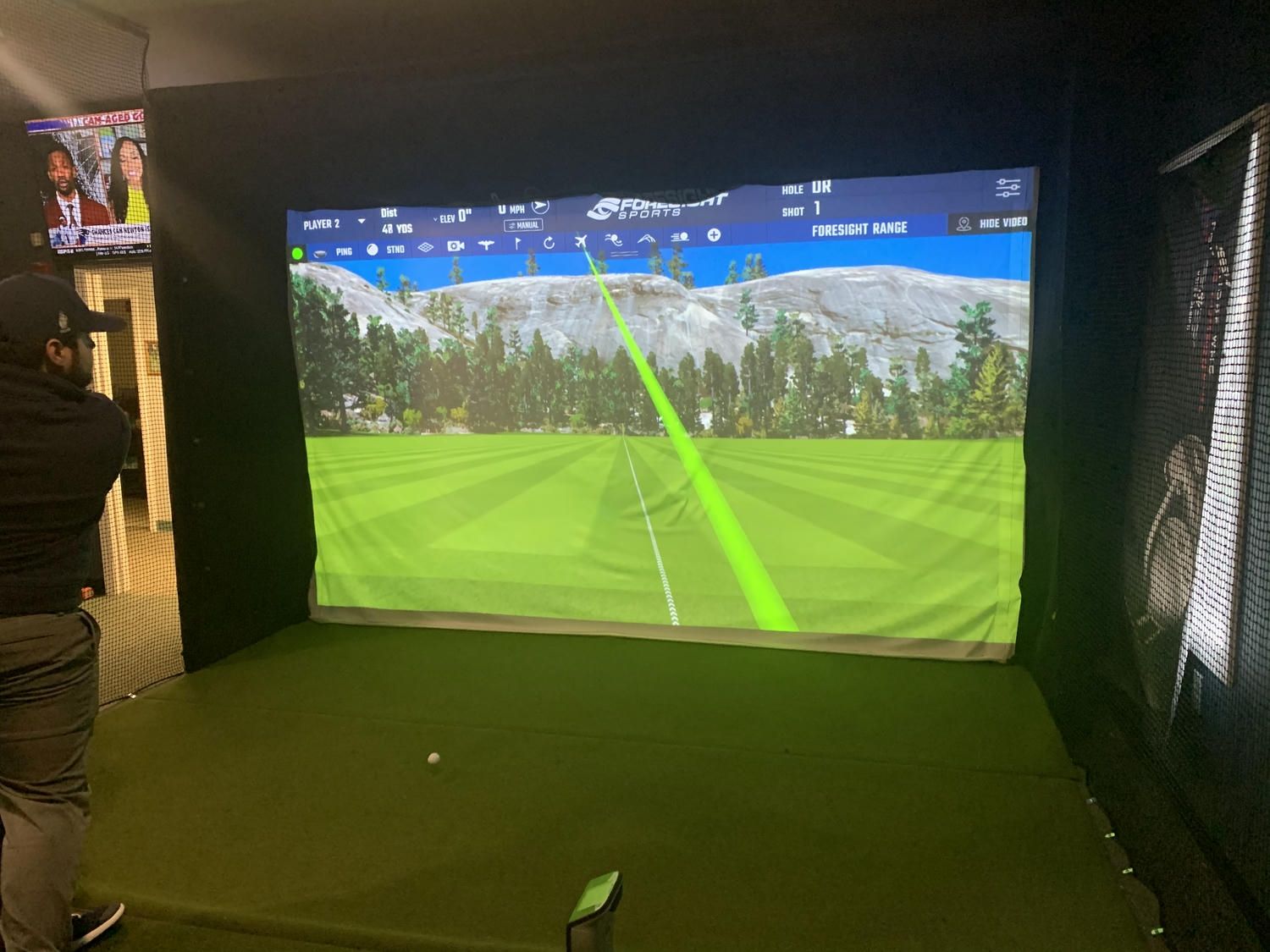 Person swings golf club in indoor simulator. Green screen displays course view, hitting a ball.