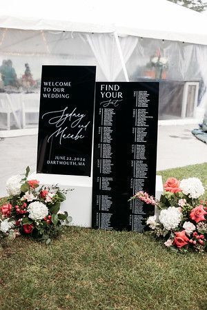 Two black signs at a wedding. One welcomes guests, the other lists seating. Flowers surround them on green grass.