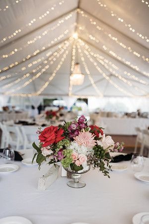 Wedding reception table with floral centerpiece. Tent with string lights, white tablecloth, and place settings.