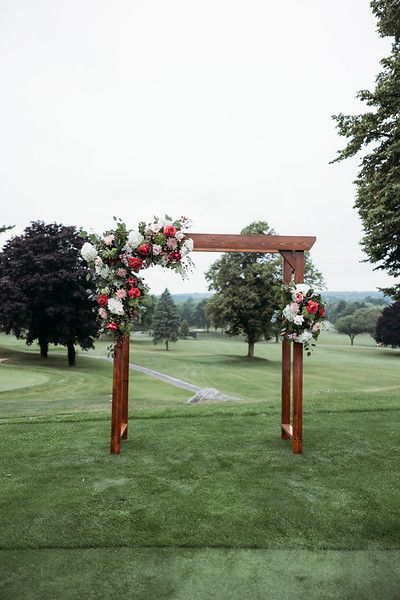 Wooden wedding arch decorated with red and white flowers on a green lawn with trees in the background.