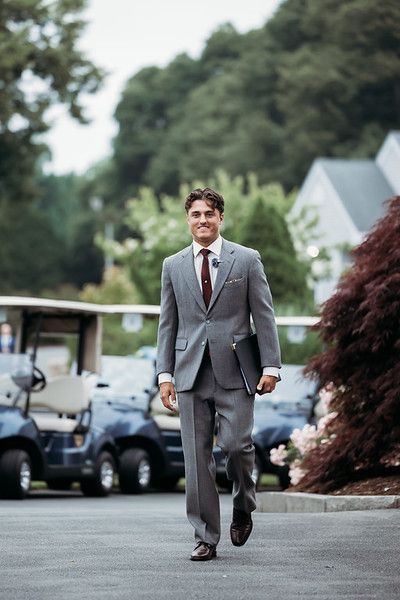 Man in gray suit walking, holding a laptop, near golf carts. Outdoors, sunny day.