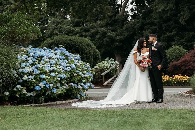 Bride and groom standing near blue hydrangeas, she in a white dress with veil, he in a black suit, looking at each other.