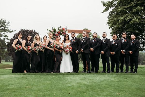 Wedding party poses on a green lawn, bride in white dress, bridesmaids in black gowns, groomsmen in black suits.