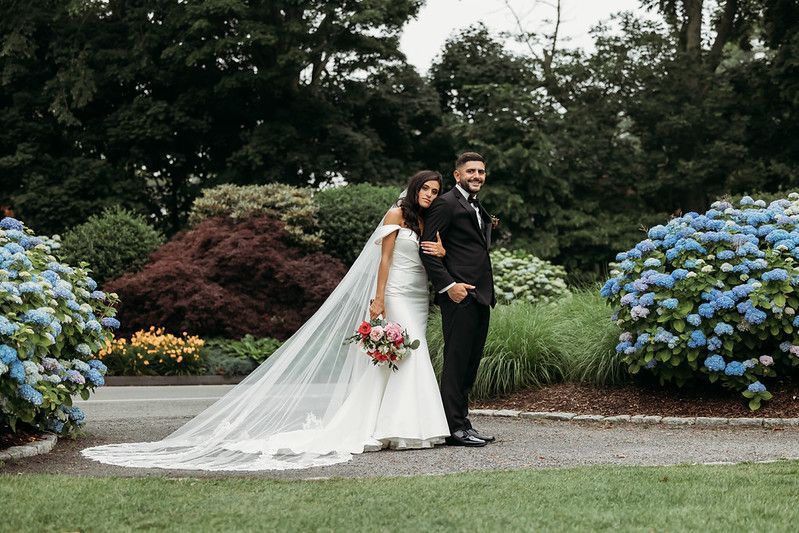 Bride and groom pose outdoors. Bride in white dress, veil. Groom in black suit. Surrounded by flowers and trees.