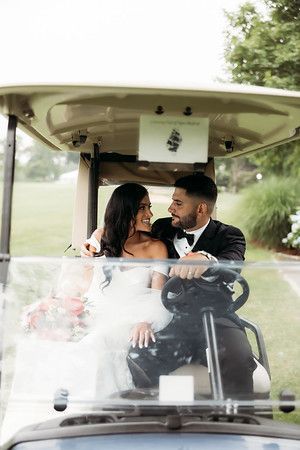 Newlyweds in a golf cart, smiling at each other. Bride in white dress, groom in a tuxedo, on a golf course.
