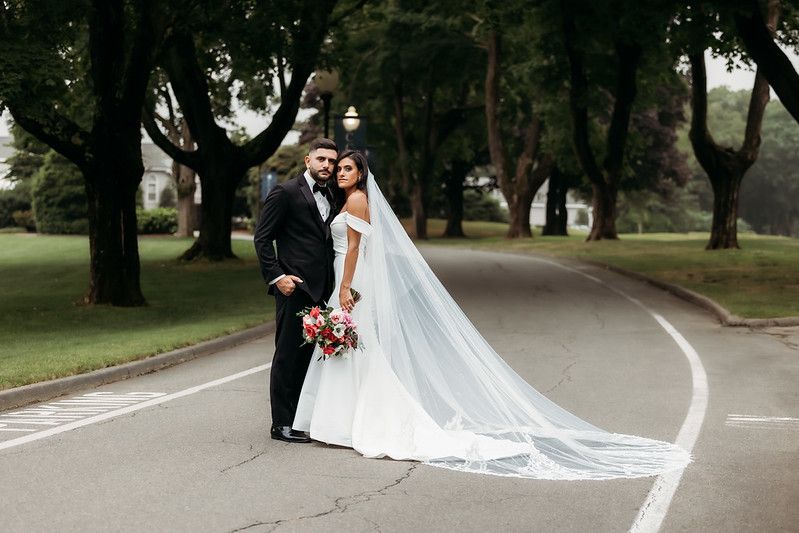 Wedding couple standing on a road, trees in the background. The bride holds a bouquet, the groom wears a black suit.