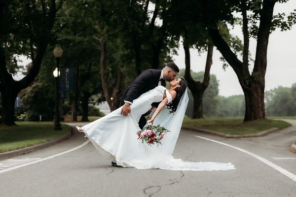 Couple embraces in a dip on an empty road. The bride wears a white gown and carries a bouquet. Trees line the sides.