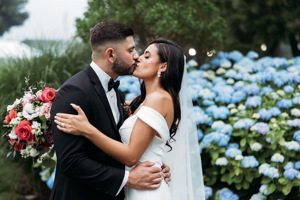 Couple kissing at wedding, surrounded by blue flowers, man in black suit, woman in white dress.