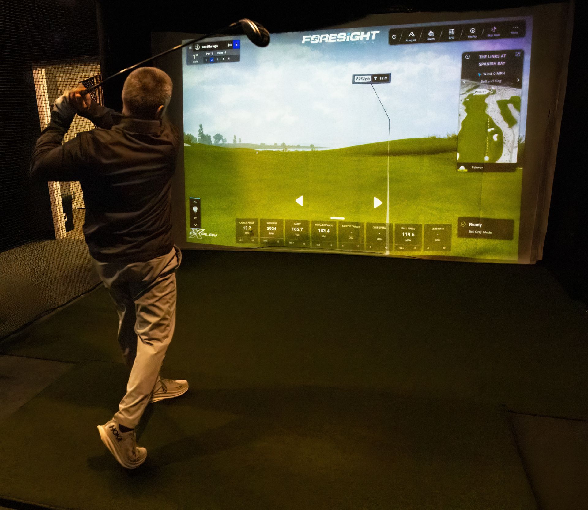 Golf simulator setup with screen displaying a golf course view. Green turf floor and black walls.