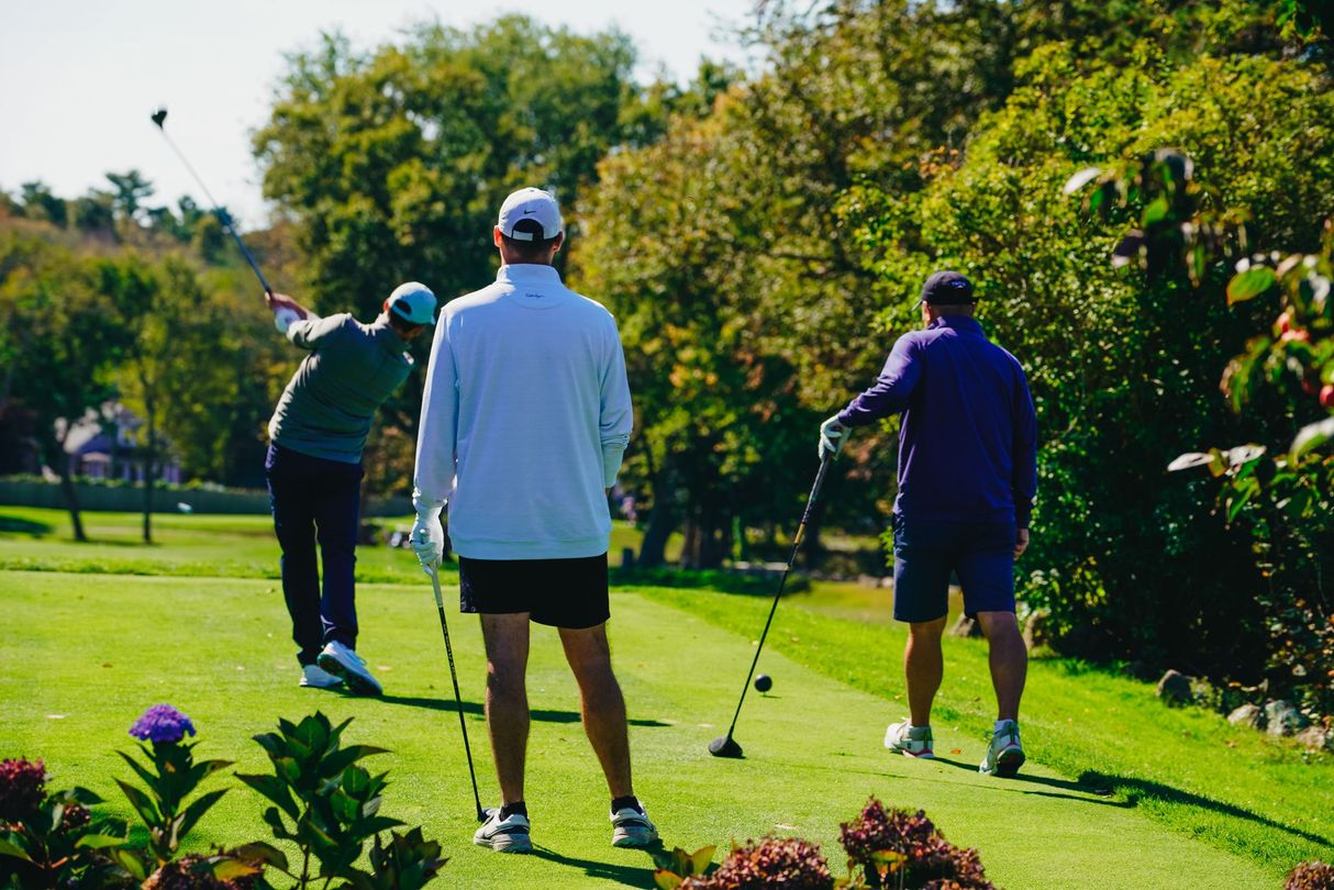 Three golfers on a green: one swinging, two watching. Sunny day with trees and a golf course.