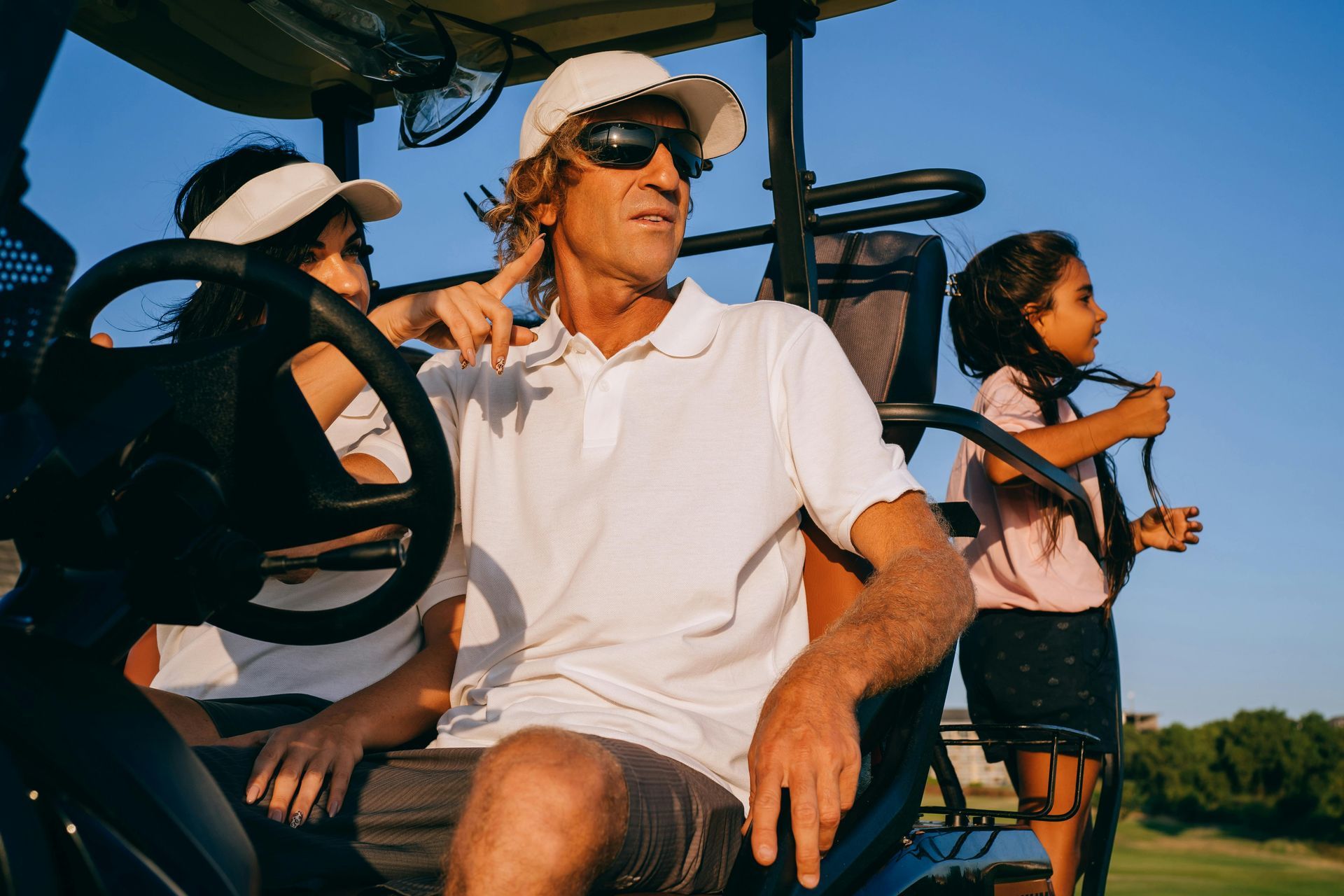Family in golf cart, man at wheel, wearing white hat and shirt, sunny outdoor setting.