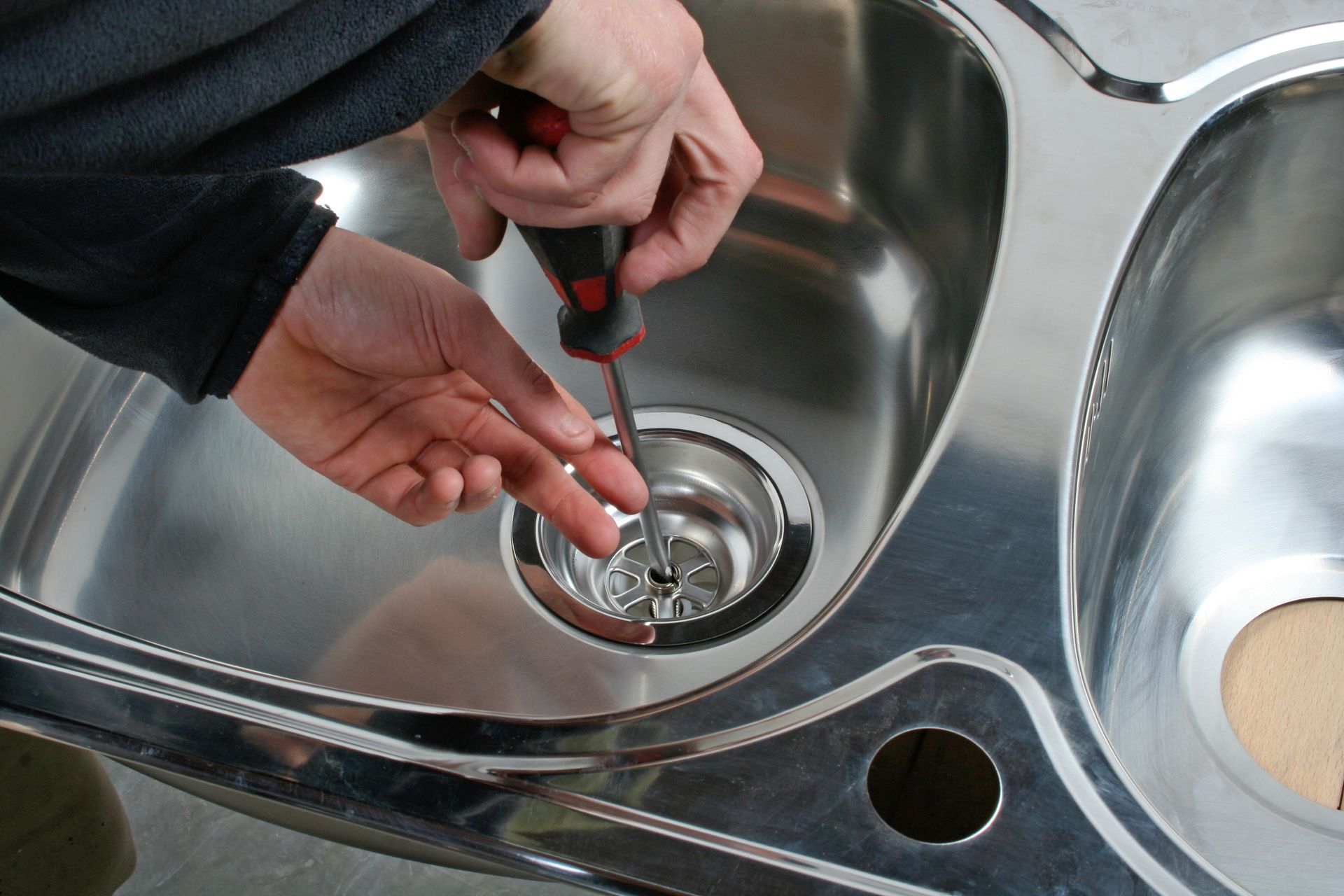 Hands using a tool to unclog a stainless steel sink drain