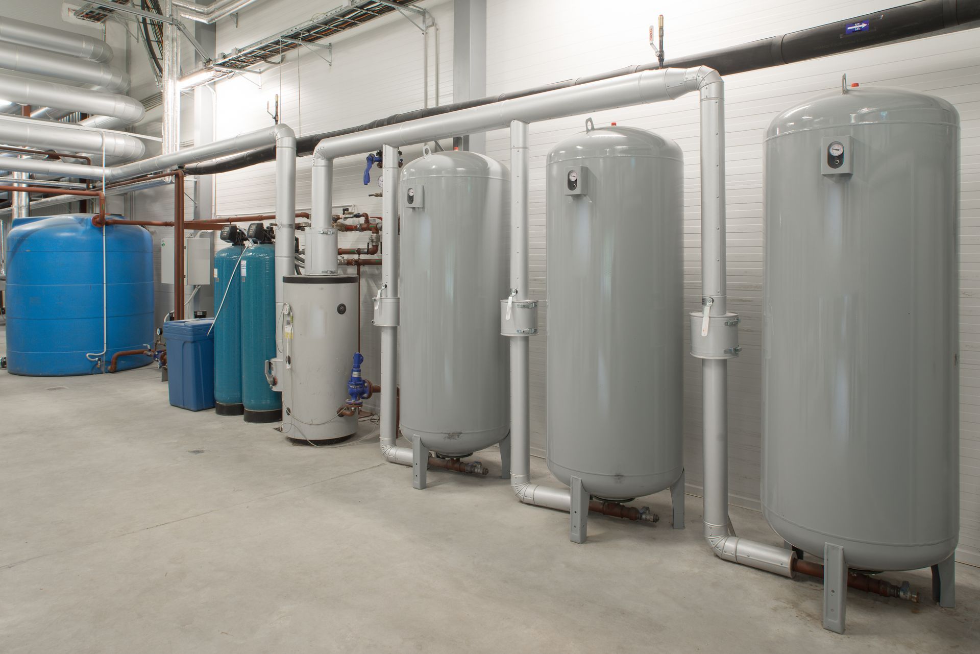 Industrial room with large gray tanks, blue barrels, and white pipes along a concrete floor