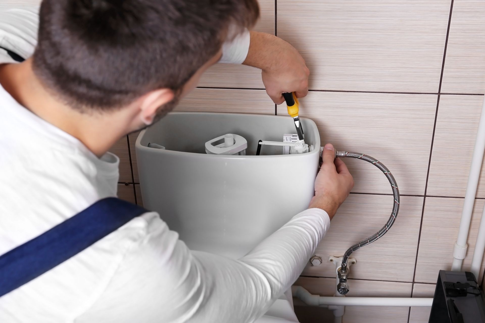 Plumber repairing a toilet tank in a tiled bathroom, with a wrench and exposed plumbing components