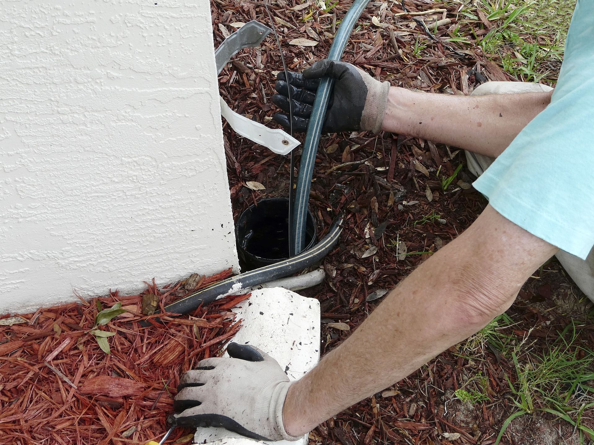 Worker using gloves to install black cable at the base of a white wall outdoors.