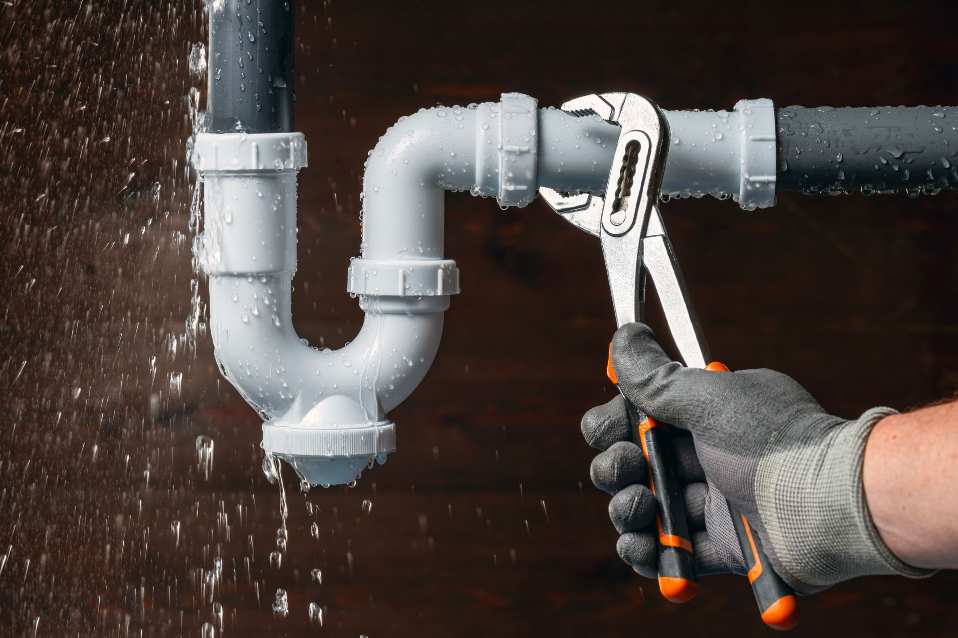 Gloved hands using a pipe wrench to fix a leaking gray drain pipe with water spraying out