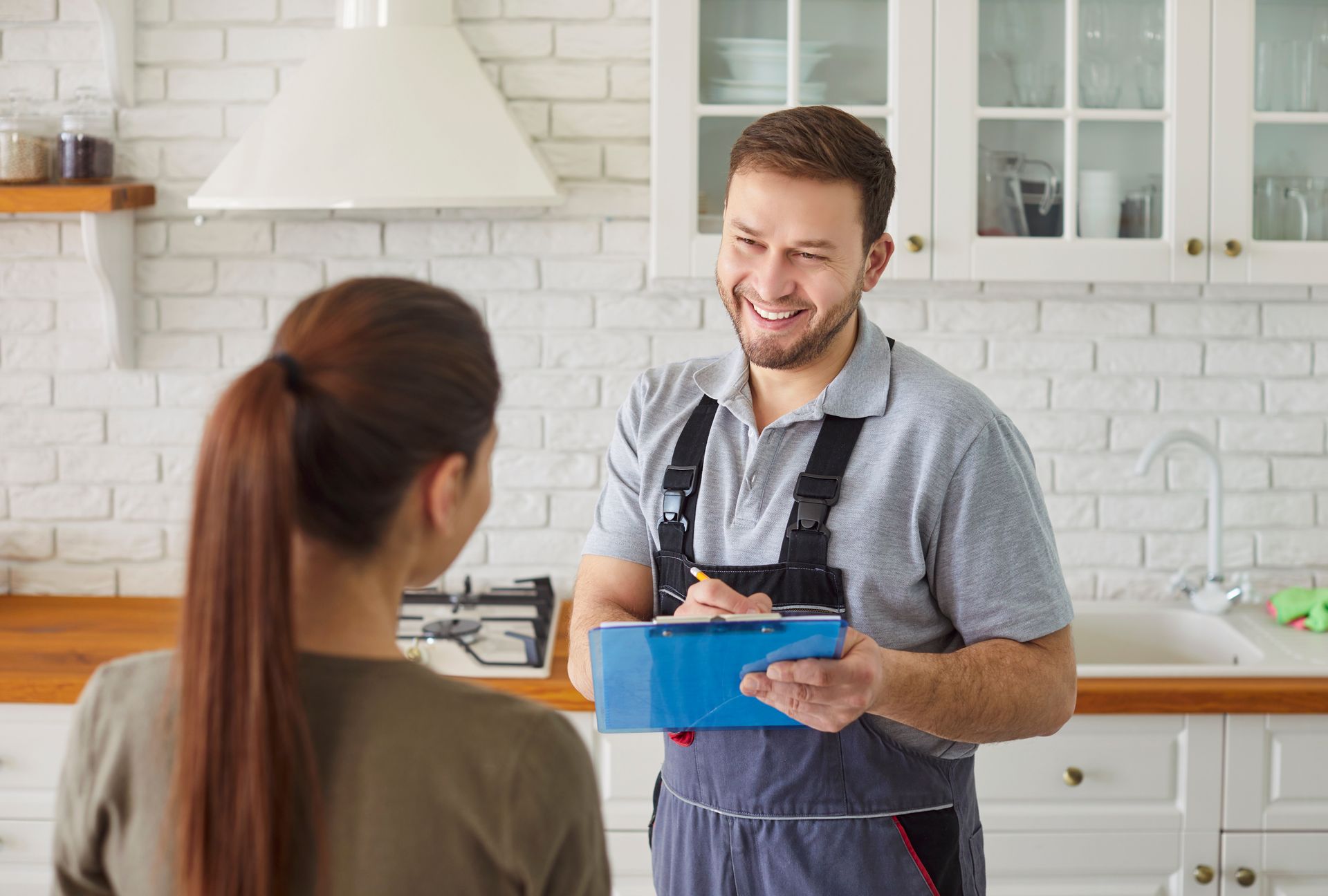 Smiling man in apron holding a blue container in a bright kitchen, talking to a woman with her back to camera