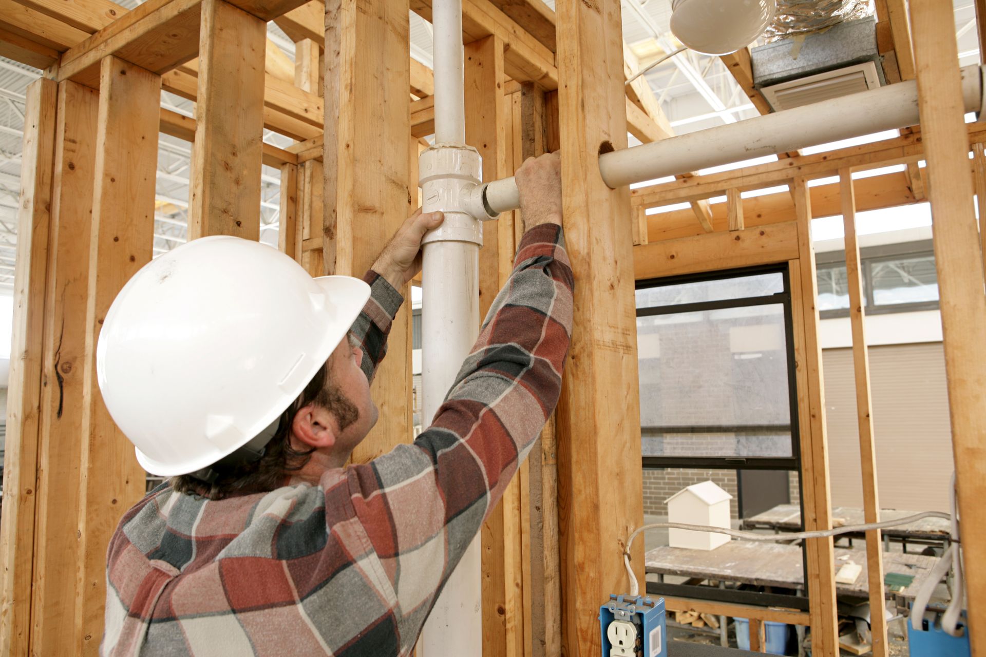 Construction worker in white hard hat installing pipes in a wooden-framed building.
