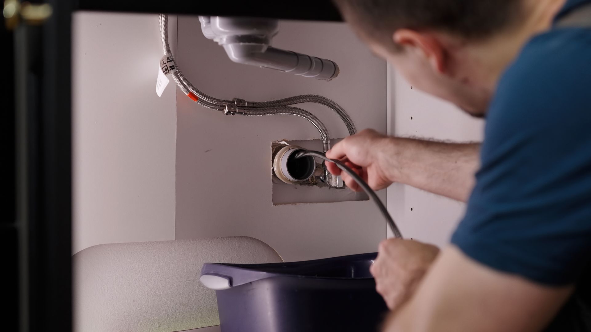 Plumber repairing a sink drain under a cabinet, holding a wrench over a bucket