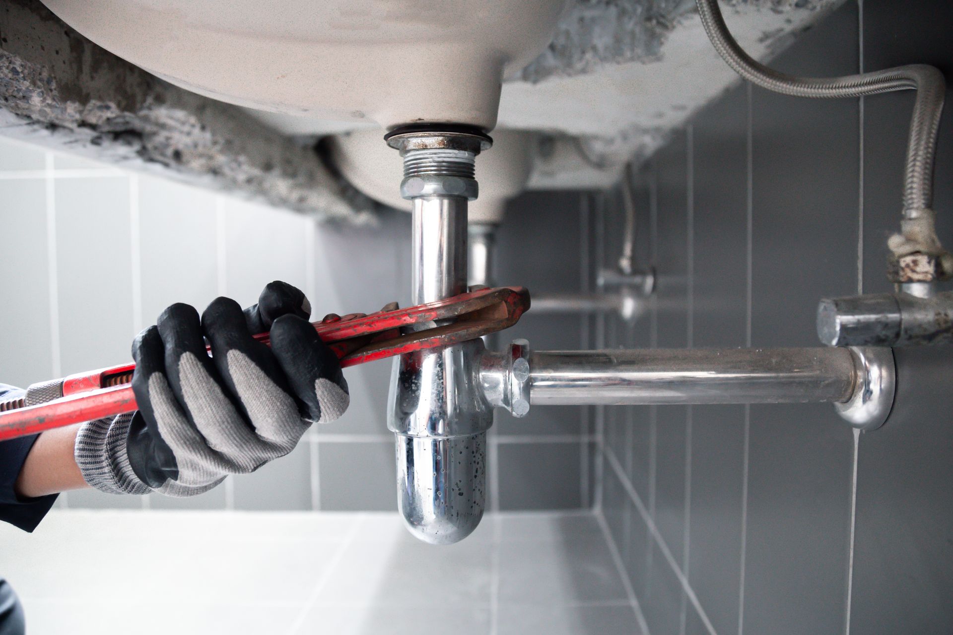Hand wearing black glove tightening a chrome sink drain pipe with a red pipe wrench under a sink