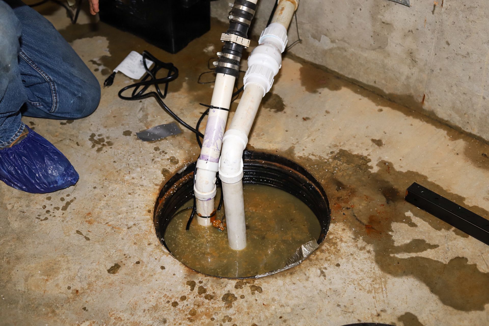 Person working on a flooded basement sump pit with PVC pipes and standing water