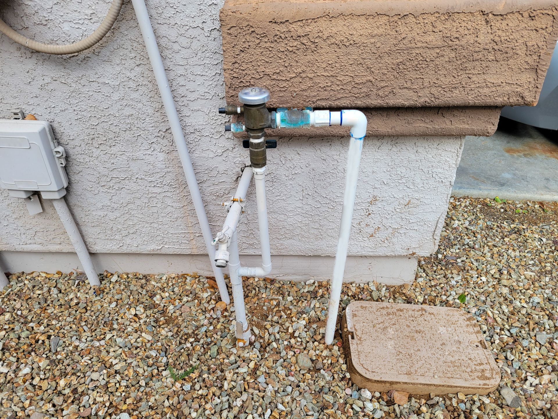 Outdoor water shutoff valve and white pipes mounted on a stucco wall beside gravel and stepping stones