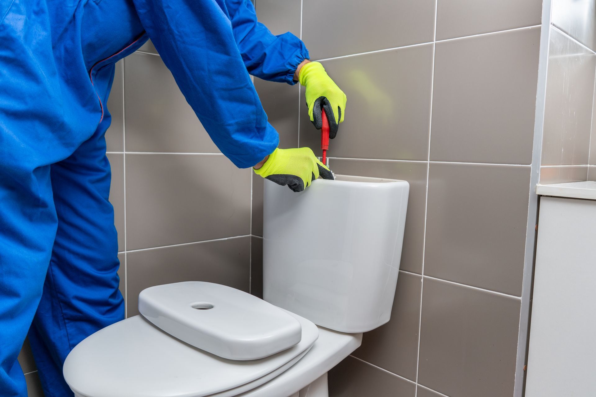 Person in blue workwear repairs a toilet flush tank with a screwdriver in a tiled bathroom.