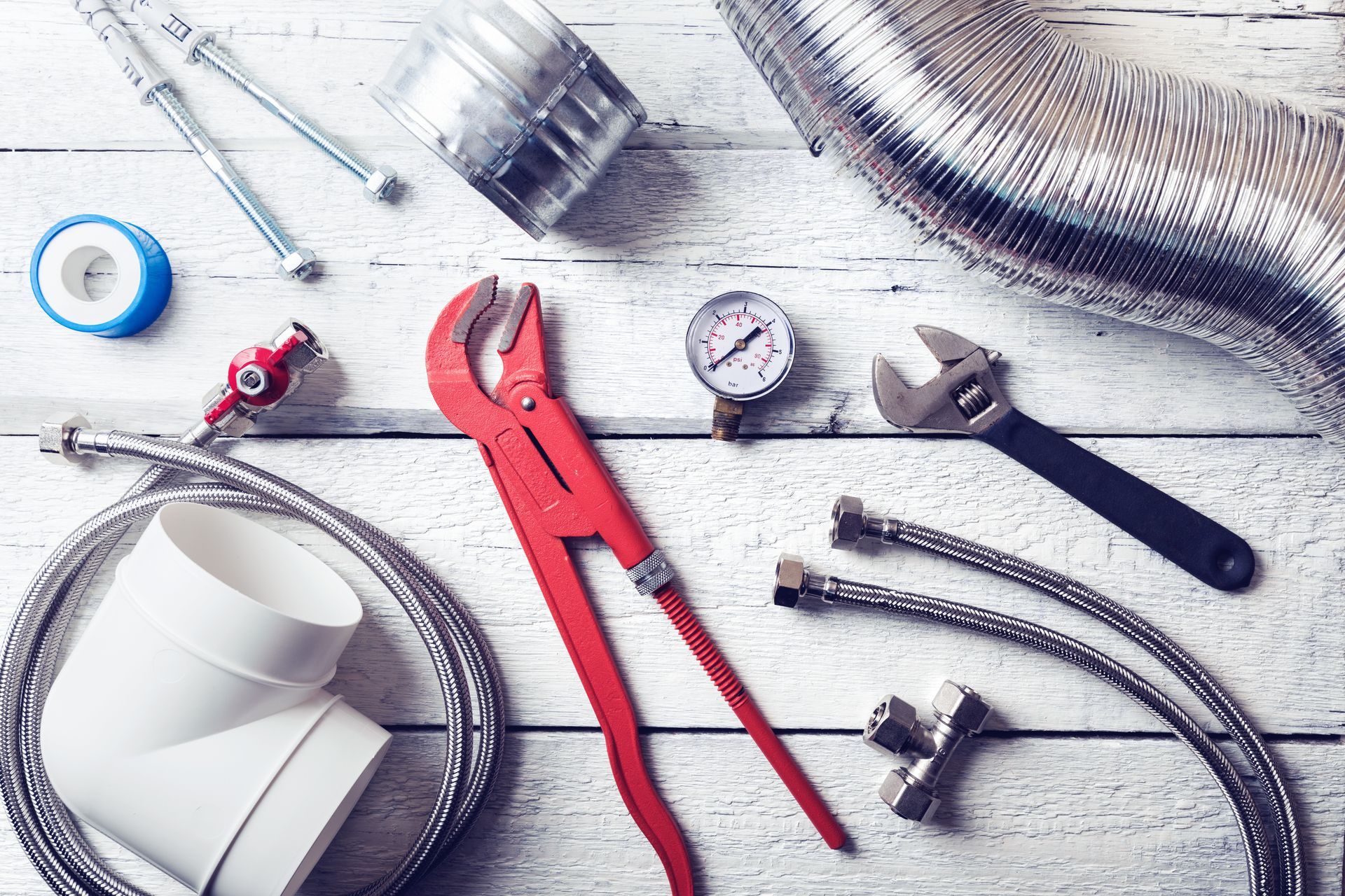 Assorted plumbing tools and hoses on a light wooden surface, including red pliers and a pressure gauge