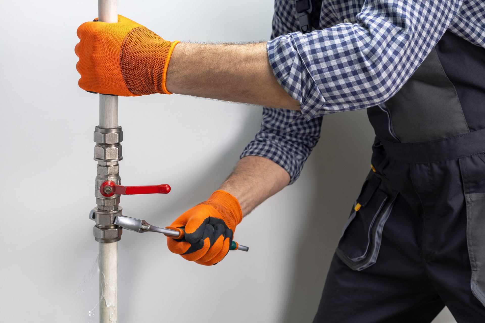 Worker tightening a pipe valve with a wrench, wearing orange gloves and checkered shirt.