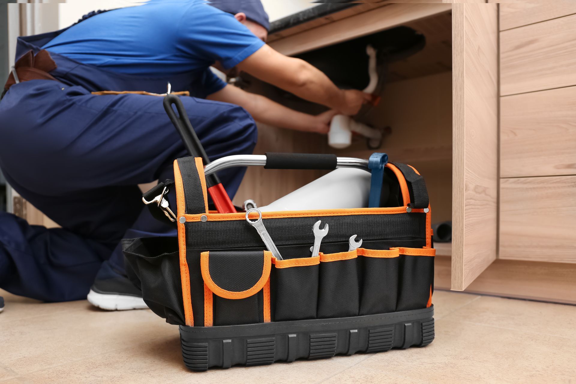 Technician kneeling under a kitchen cabinet beside a black-and-orange tool bag
