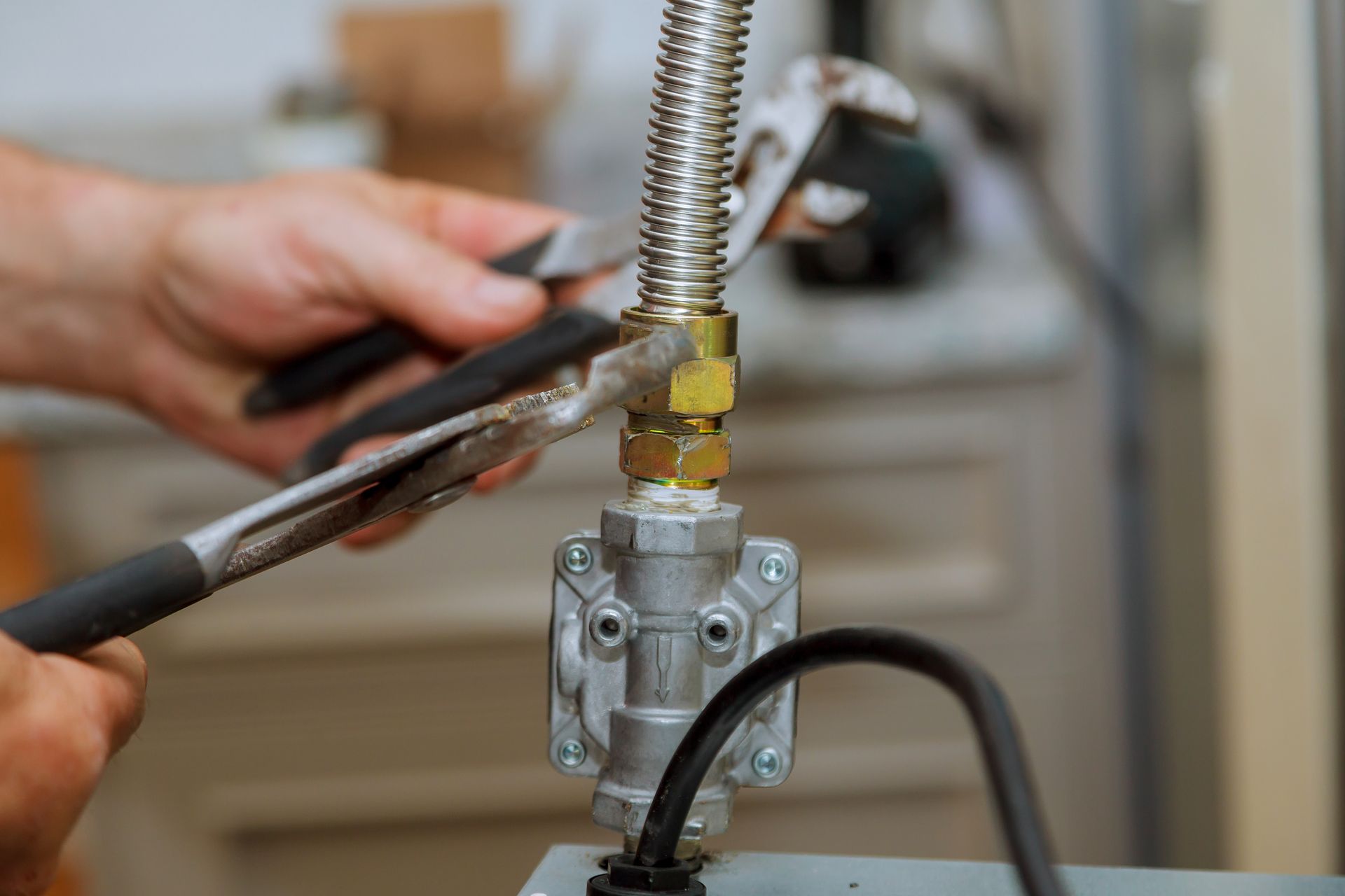 Hands using pliers to work on a metal pipe valve with a flexible hose attachment.