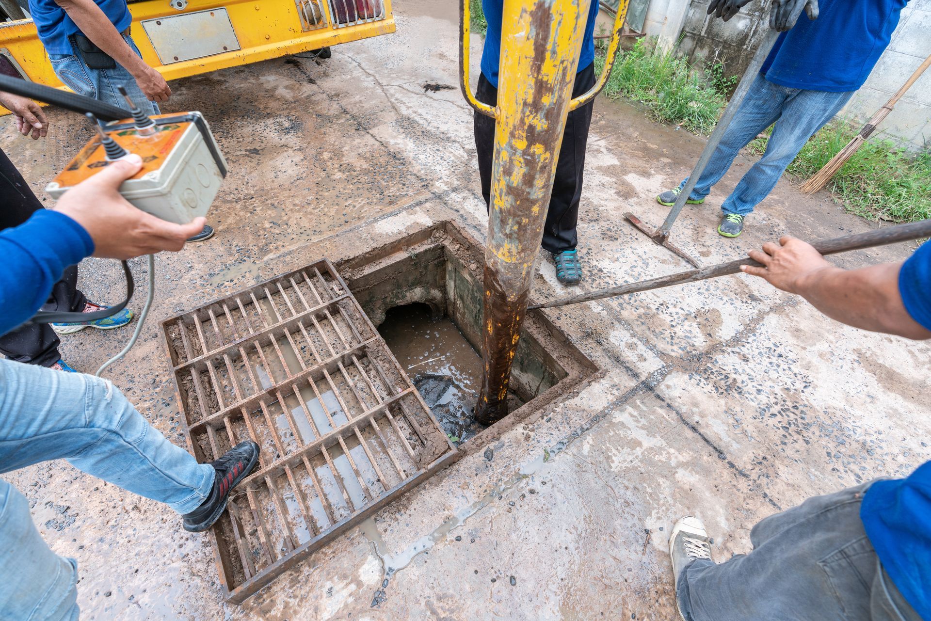 Workers removing a metal grate from a concrete pit beside yellow machinery, with tools and hands visible